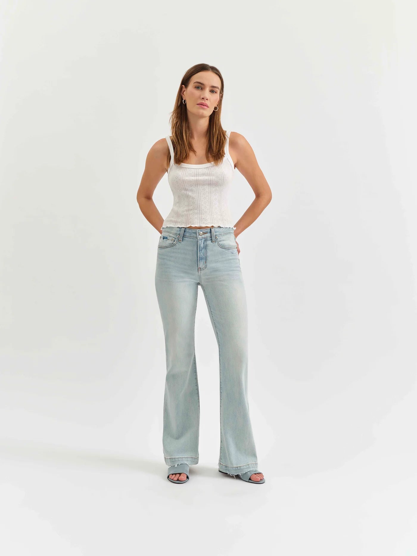 Model wearing the mid-rise flare jeans by Daze Denim in swoon vintage wash. Paired with a white tank top and blue slides.