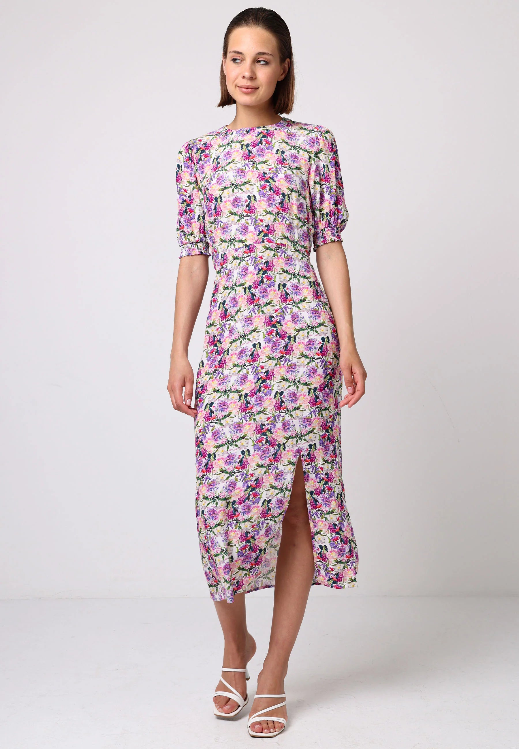 Model wearing Angeleye's pink floral midi dress with puff sleeves, elastic waist, and front slit. Styled with white strappy heels against a light studio backdrop.