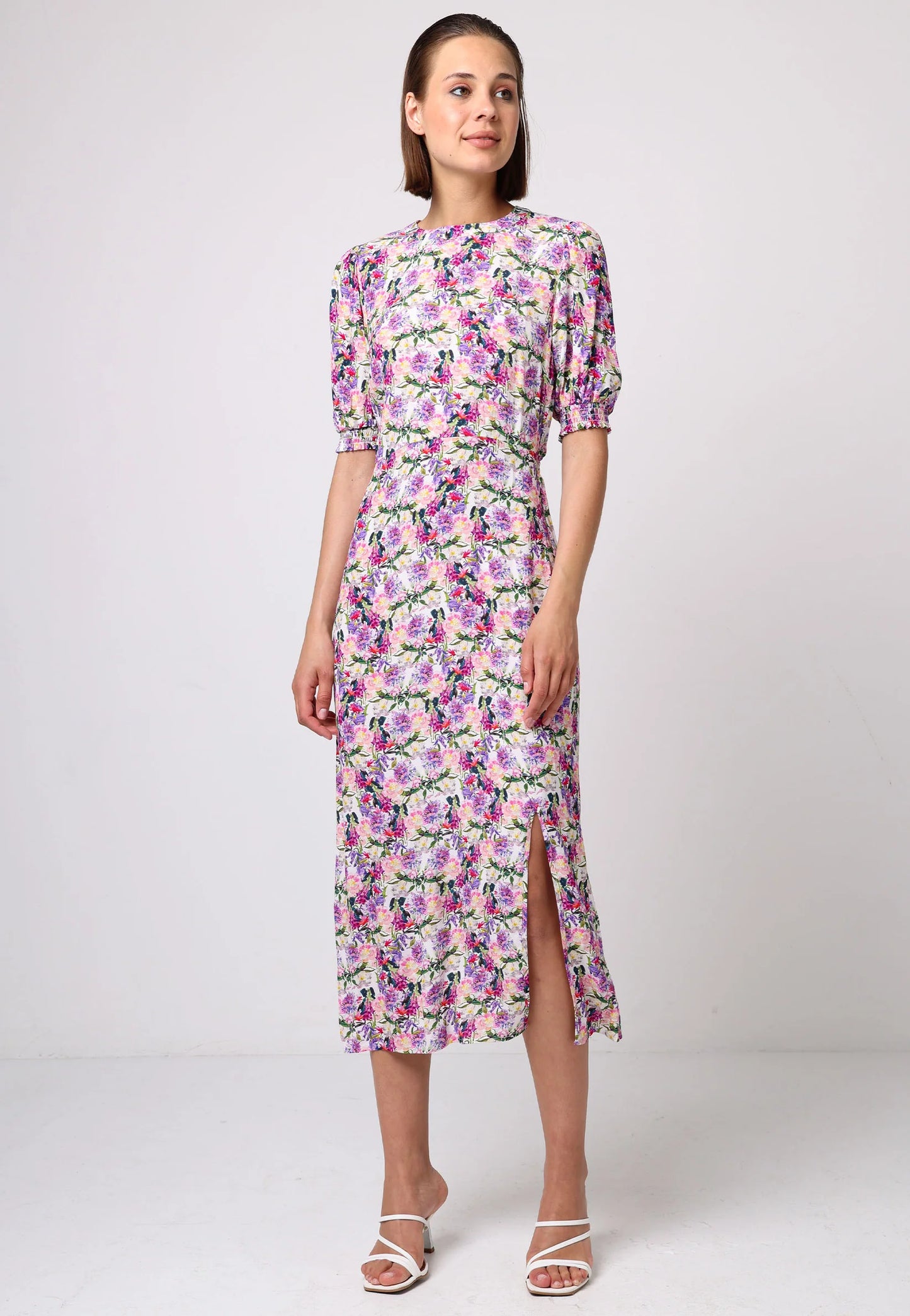 Model wearing pink floral midi dress with puff sleeves, elastic waist, and front slit. Styled with white strappy heels against a light studio backdrop.