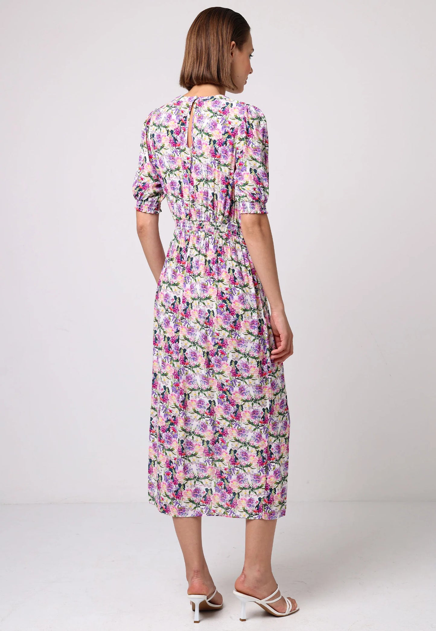 Back view of a model wearing pink floral midi dress with puff sleeves, elastic waist, and front slit. Styled with white strappy heels against a light studio backdrop.