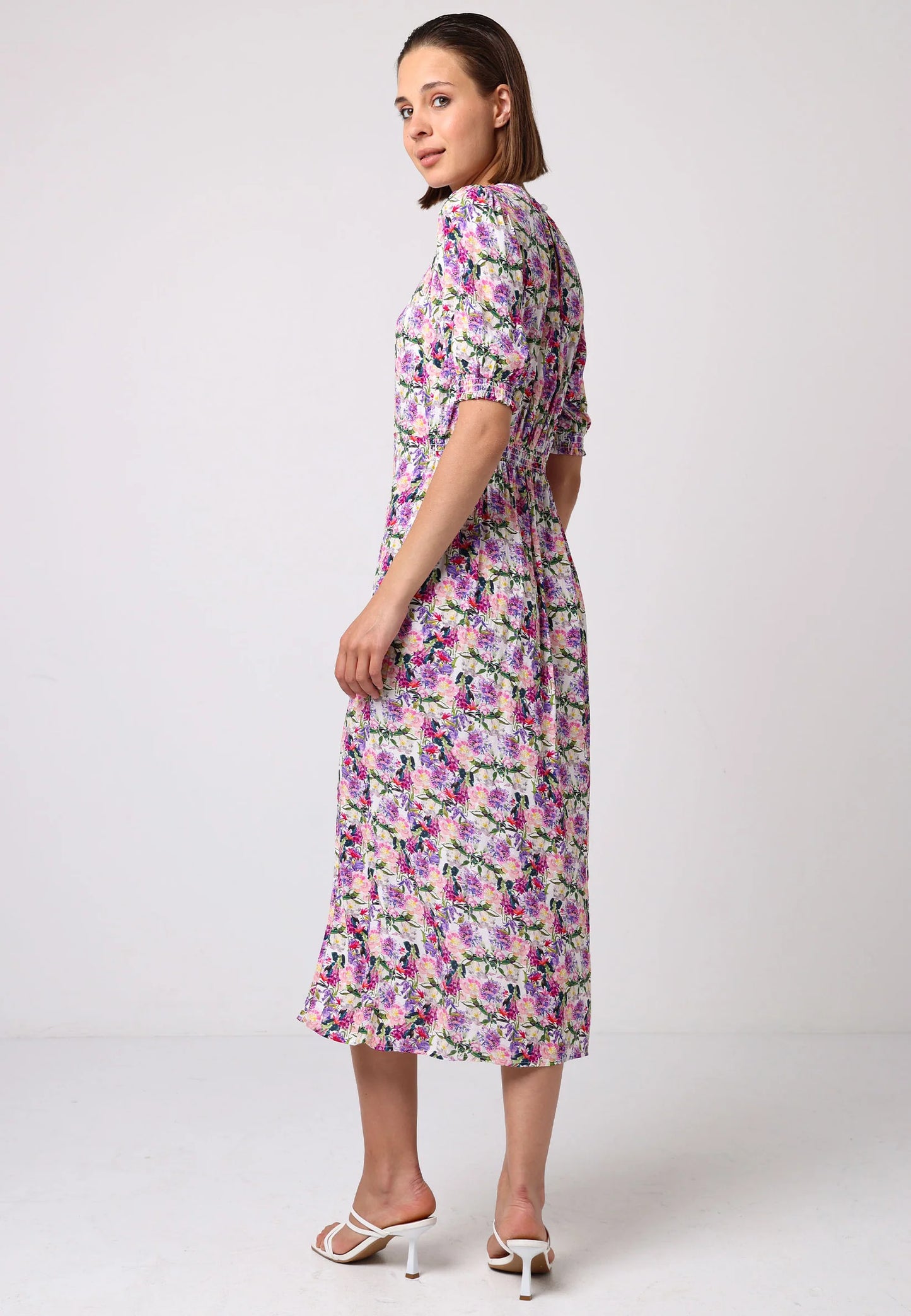 Side/back view of model wearing pink floral midi dress with puff sleeves, elastic waist, and front slit. Styled with white strappy heels against a light studio backdrop.