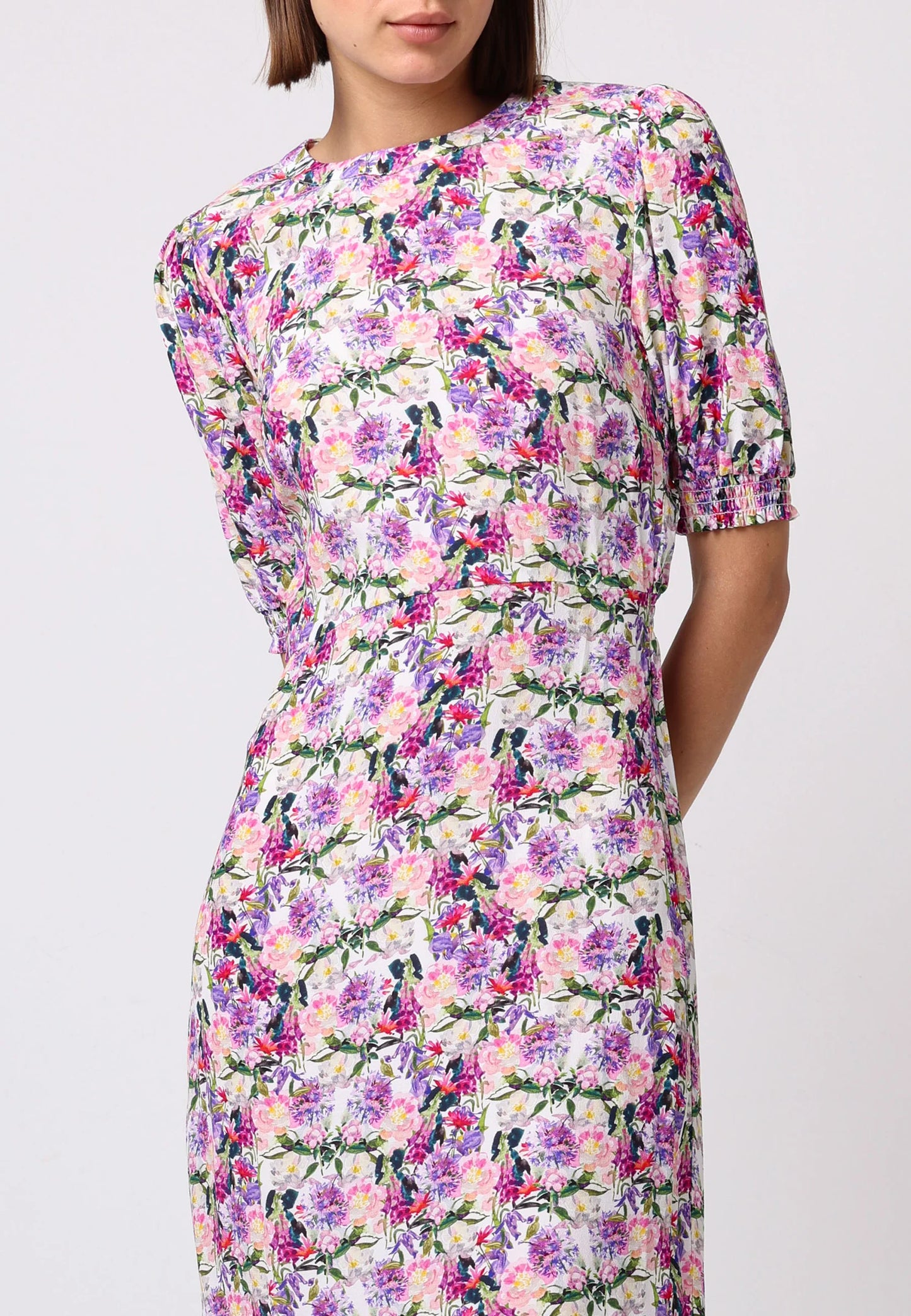 Close up view of model wearing pink floral midi dress with puff sleeves, elastic waist, and front slit. Styled with white strappy heels against a light studio backdrop.