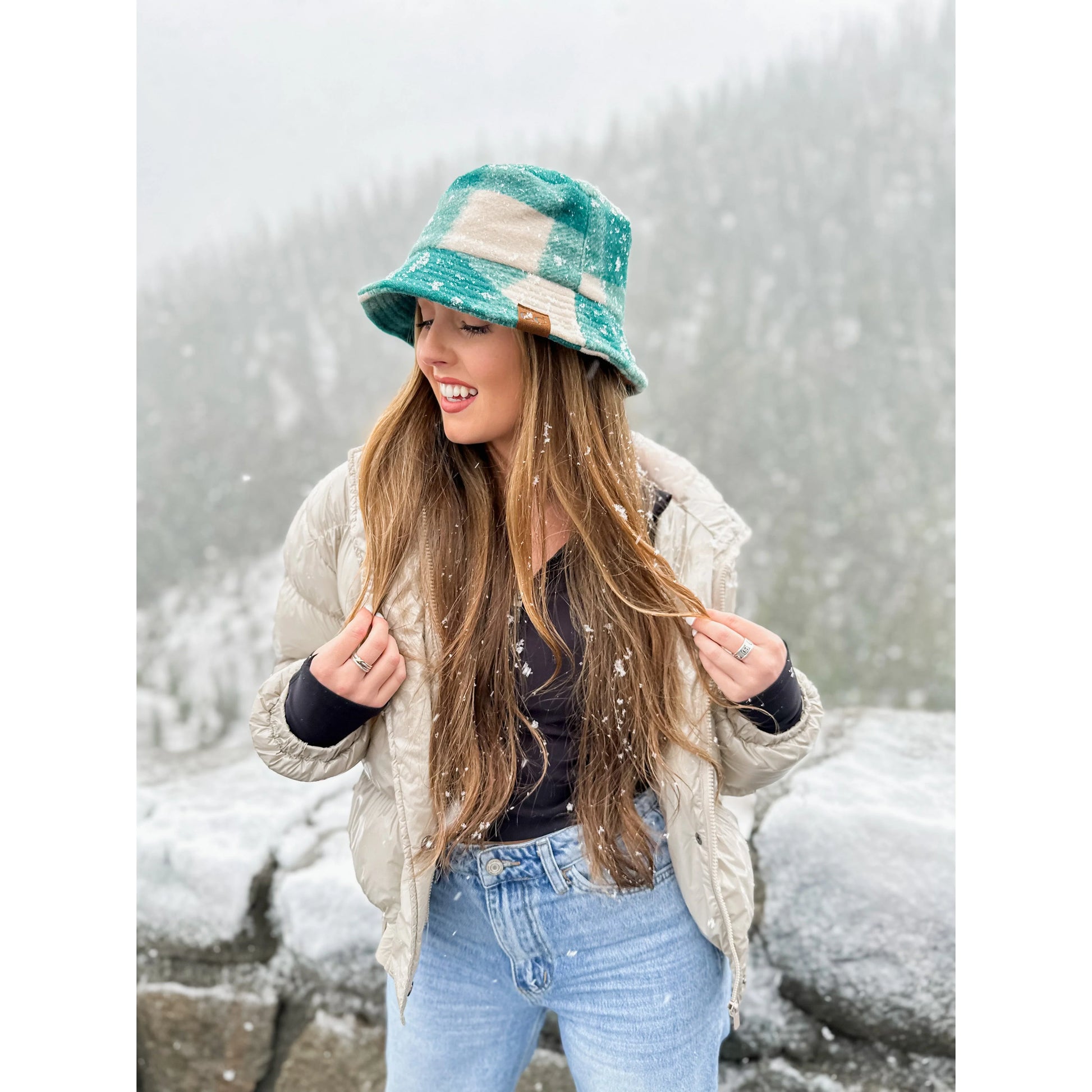 Model wearing the C.C Beanie Plaid Bucket Hat in green and tan, standing in a snowy outdoor setting with a beige puffer jacket and jeans.