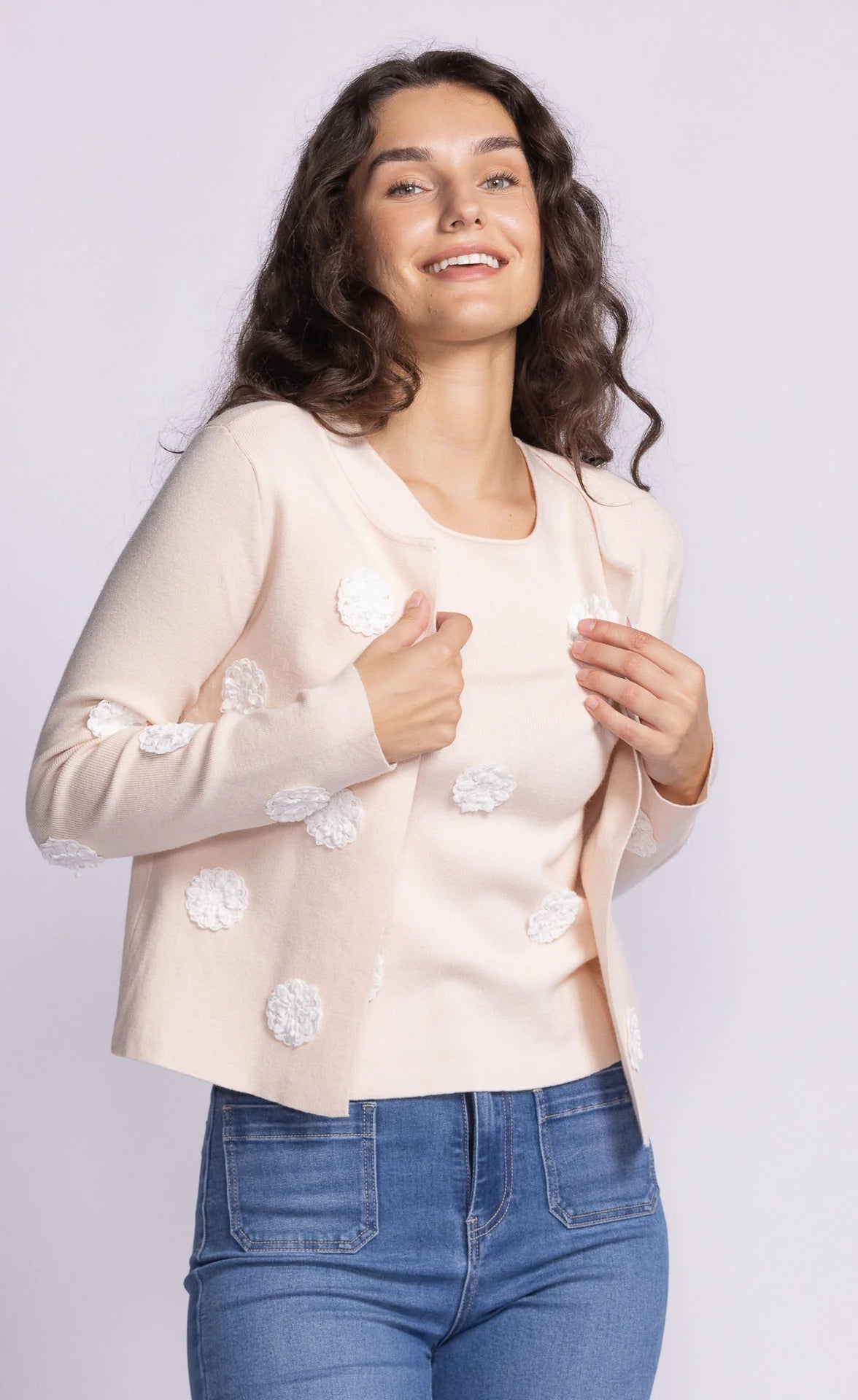 Brunette Model wearing Pink Martini's Kelsey Sweater in light peach, featuring a delicate open-front design with 3D floral appliqué detailing.