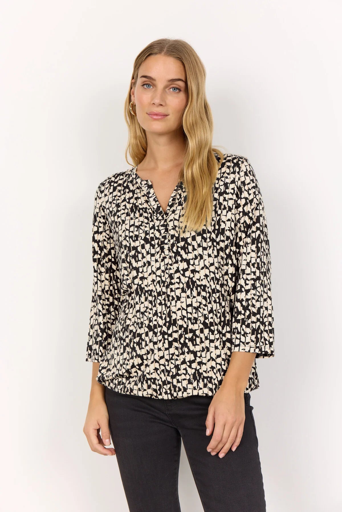 A model wearing the Soya Concept Marica AOP 316 3/4 Sleeve Top in Black Beige Print. The top features a relaxed fit, a subtle V-neckline, and a stylish abstract pattern. She pairs it with black pants and stands with one hand in her pocket. Model wears size M.