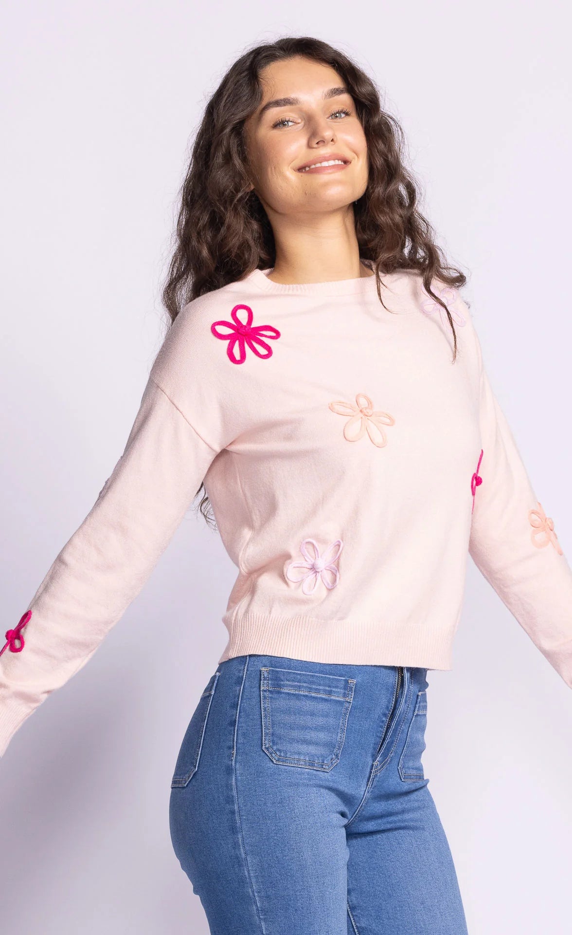 Front view of the Pink Martini Evie Sweater in light pink, featuring colorful floral appliqués and a relaxed fit, styled with denim jeans.