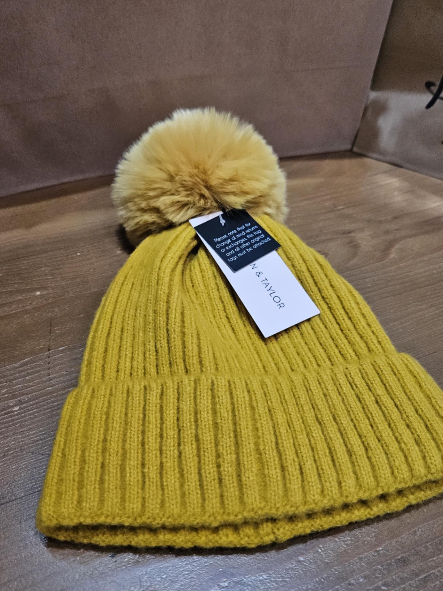 Golden yellow Morgan Taylor Lula Beanie with a pom-pom, displayed flat on a wooden surface with its tag visible.