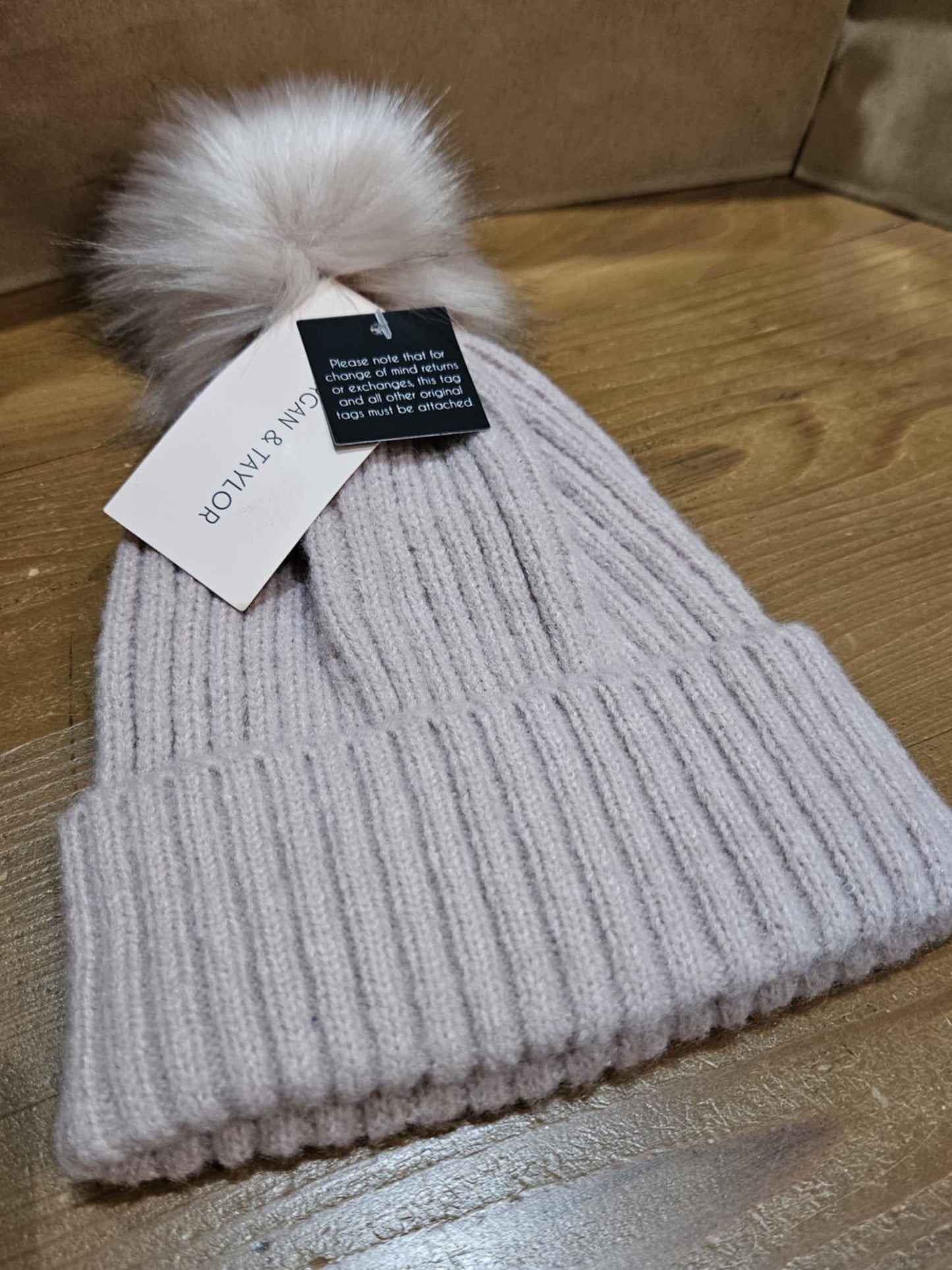 Pale pink Morgan & Taylor Zara Beanie with a faux fur pom-pom, laying flat on a wooden surface, showcasing its ribbed knit texture and cozy design.