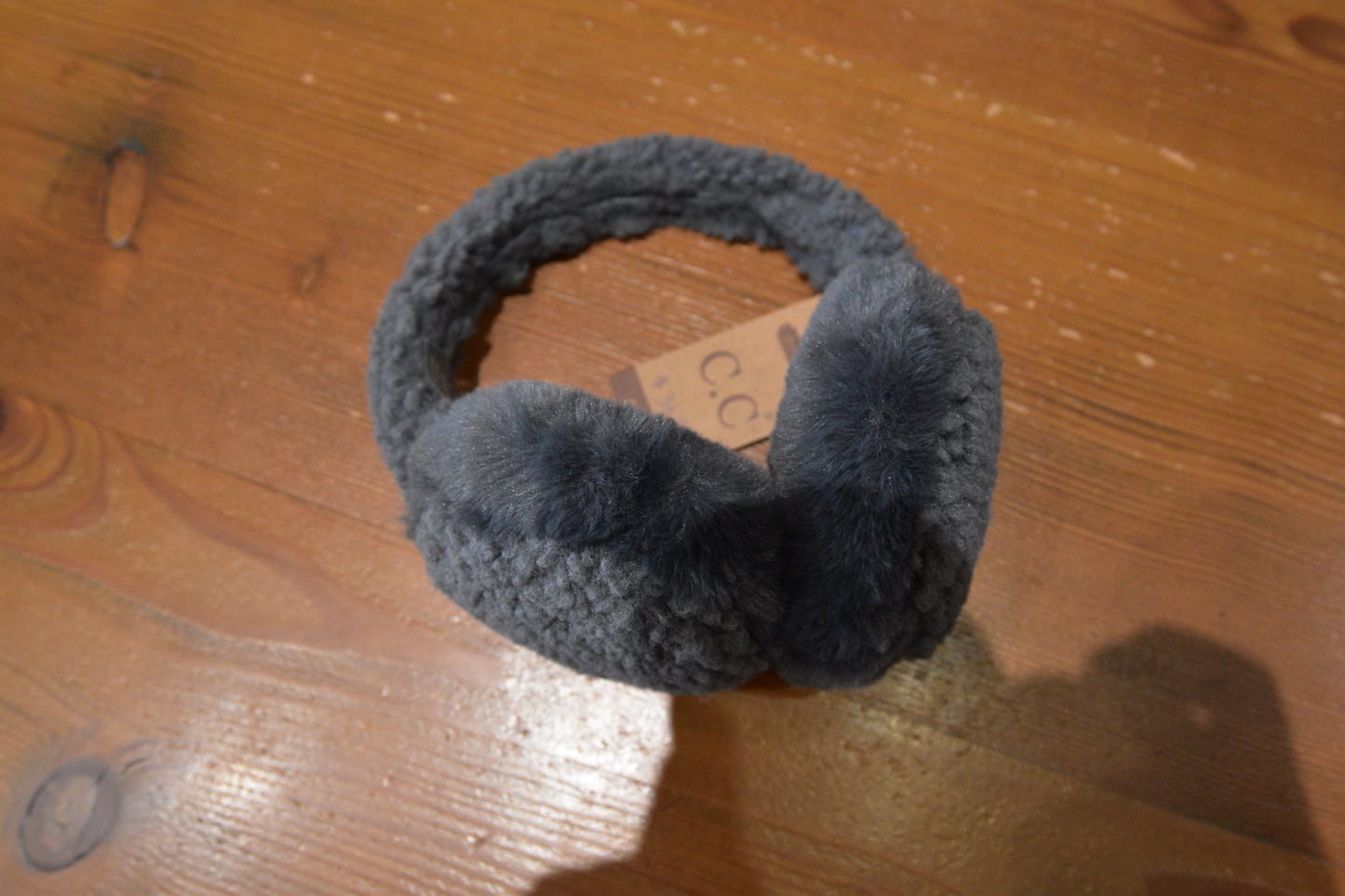 Close-up of C.C Beanie Faux Fur Sherpa Earmuffs in grey, displayed on a wooden surface, highlighting the luxurious faux fur and Sherpa-lined headband.