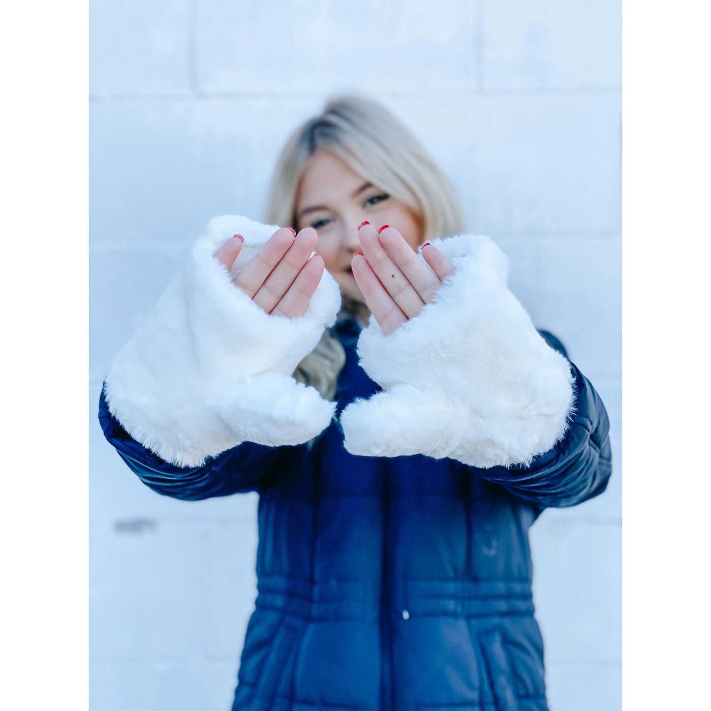 Model wearing the white fuzzy convertible mittens, holding her hands up outdoors, showcasing their stylish and cozy look with a blue winter coat. 