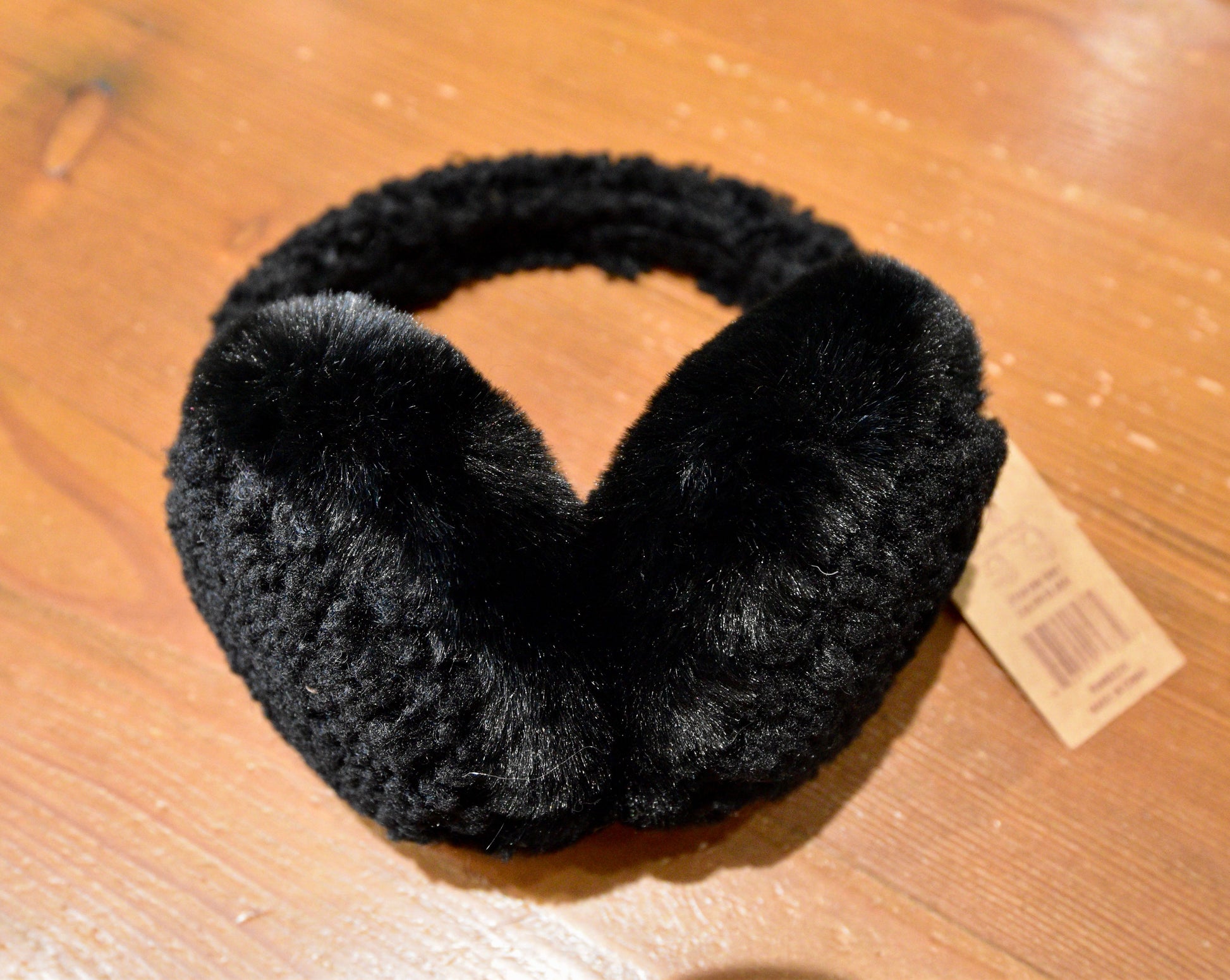 Close-up of C.C Beanie Faux Fur Sherpa Earmuffs in black, displayed on a wooden surface, showcasing their plush ear covers and soft Sherpa-lined band.