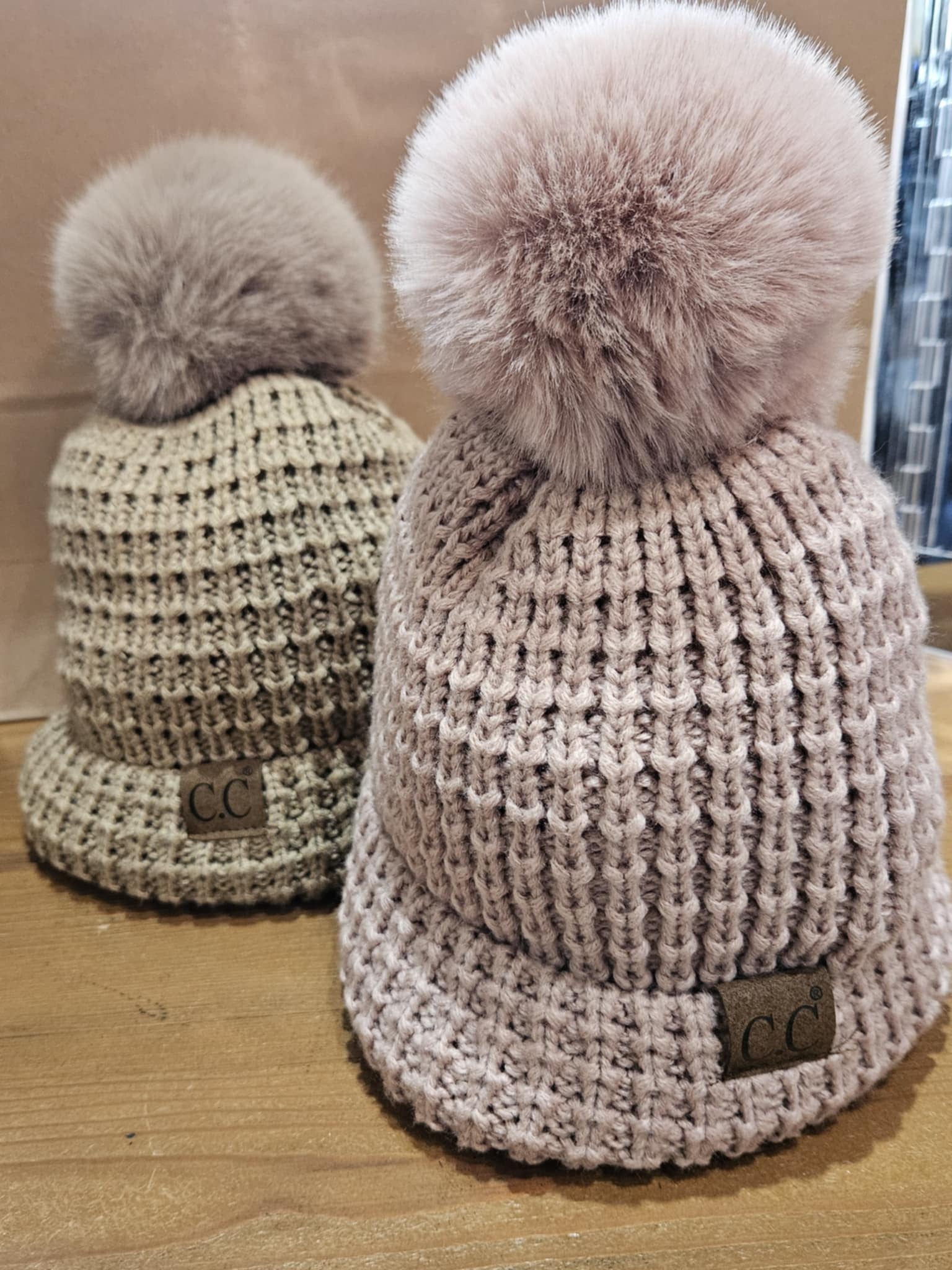 Close-up of two C.C Beanie Knit Pom-Pom Hats in tan and dusty pink, folded and displayed on a wooden surface, showcasing their leather logo tags.