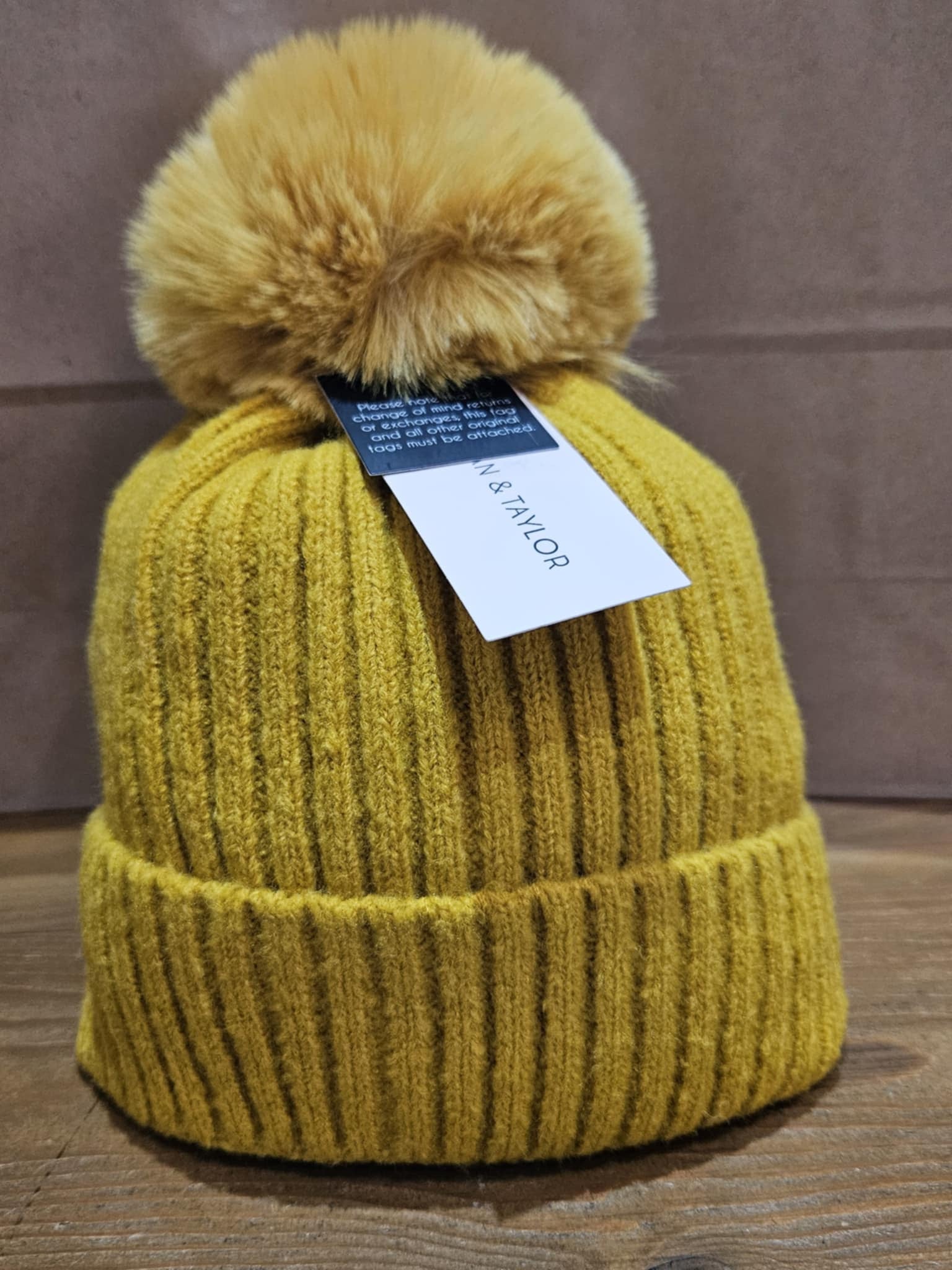 Front view of the golden yellow Morgan Taylor Lula Beanie, showing its ribbed texture and fluffy pom-pom on a wooden surface.