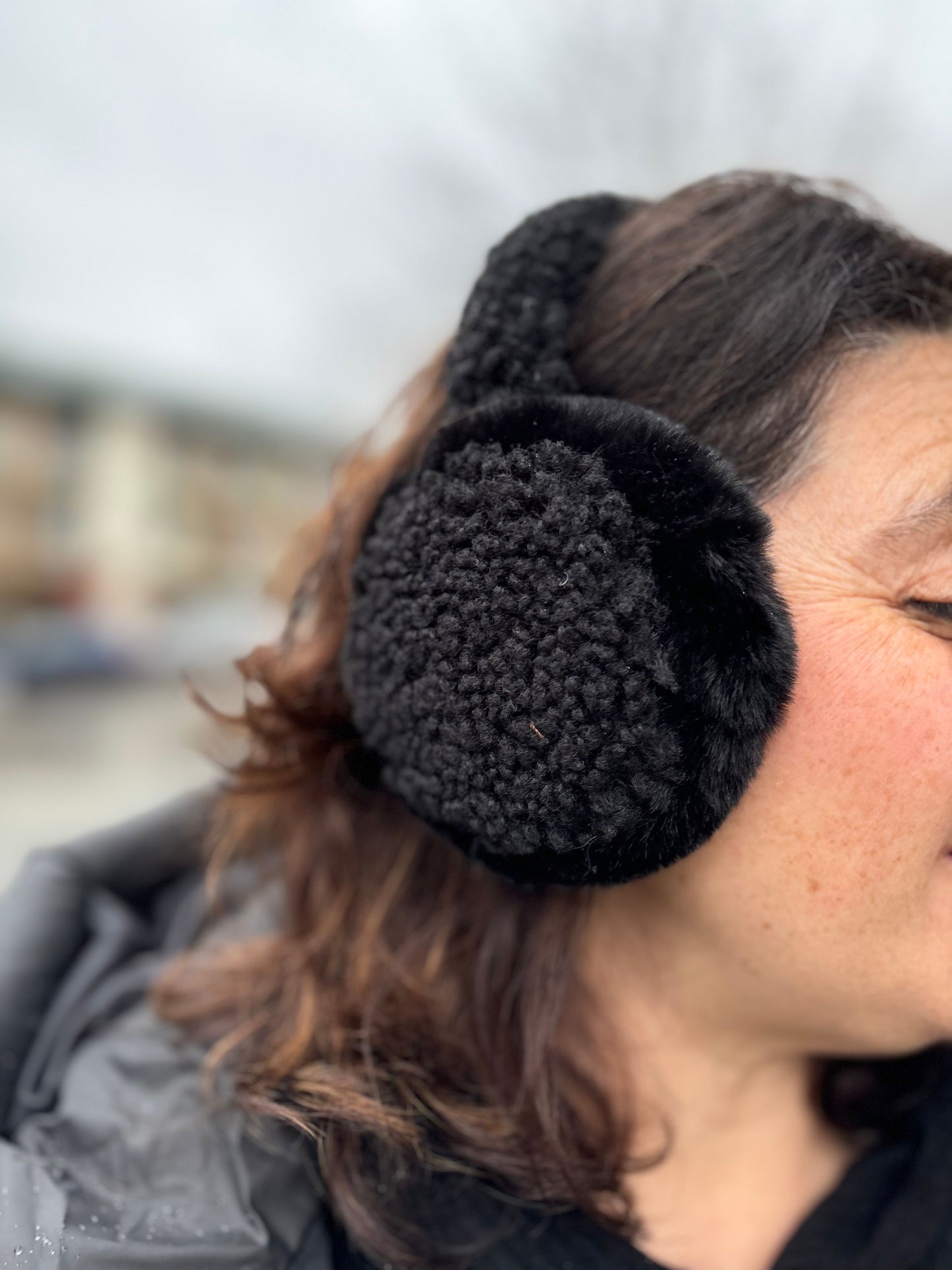 Side view of a model wearing grey C.C Beanie Faux Fur Sherpa Earmuffs, standing outside on a cold day with a matching winter jacket.