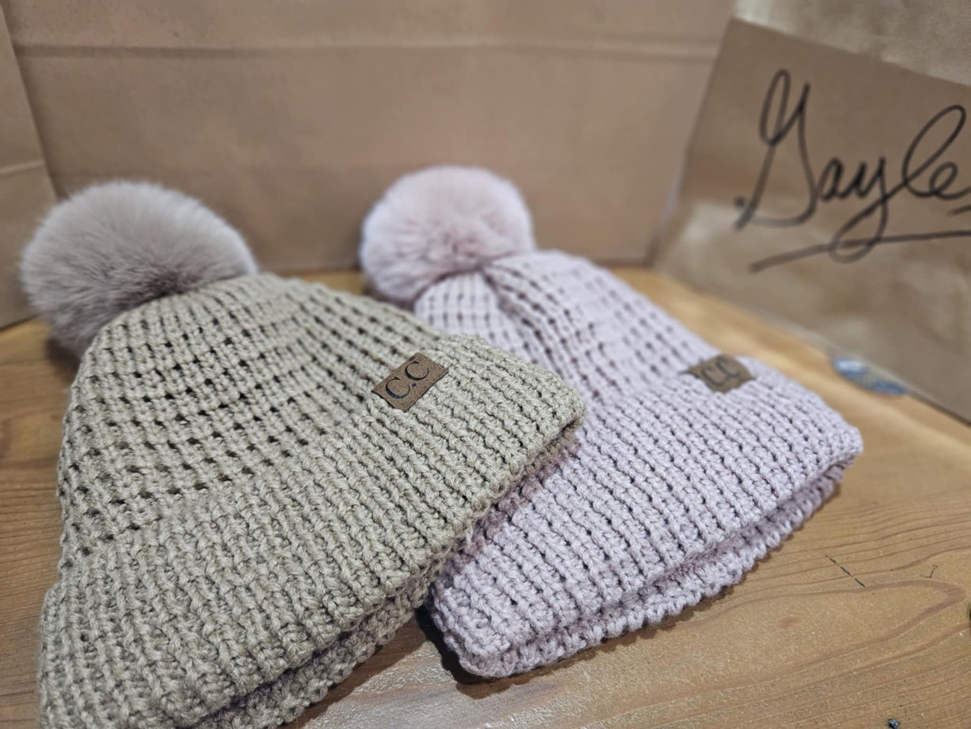 Pair of C.C Beanie Knit Pom-Pom Hats in dusty pink and tan, displayed side by side on a wooden surface, highlighting their cozy knit and soft pom-poms.