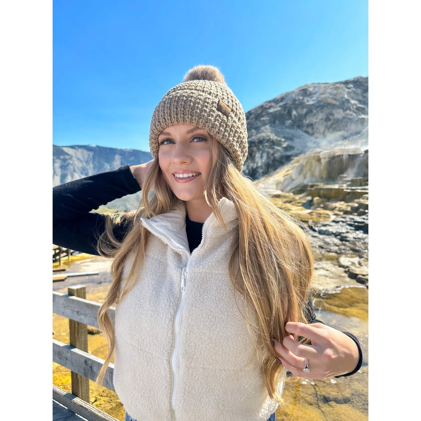 Model wearing the tan C.C Beanie Knit Pom-Pom Hat in a mountain setting, styled with a white fleece vest and long blonde hair flowing.