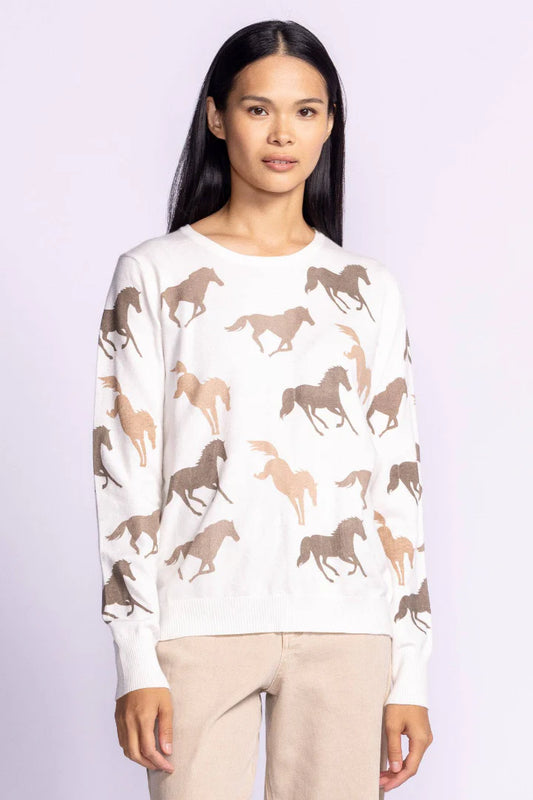 Front view of the Wild Horses sweater by Pink Martini, a cozy white knit pullover with a brown horse print, styled with beige pants and worn by a model with long dark hair.