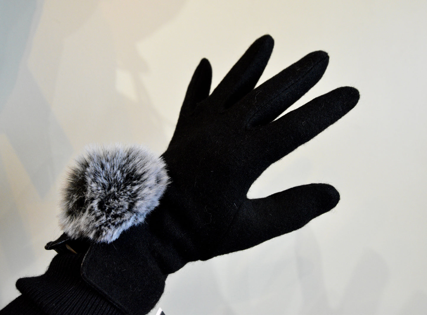 Model wearing Morgan & Taylor Antonella Gloves, lifting one hand to show the soft grey pom-pom detail in a well-lit indoor setting.