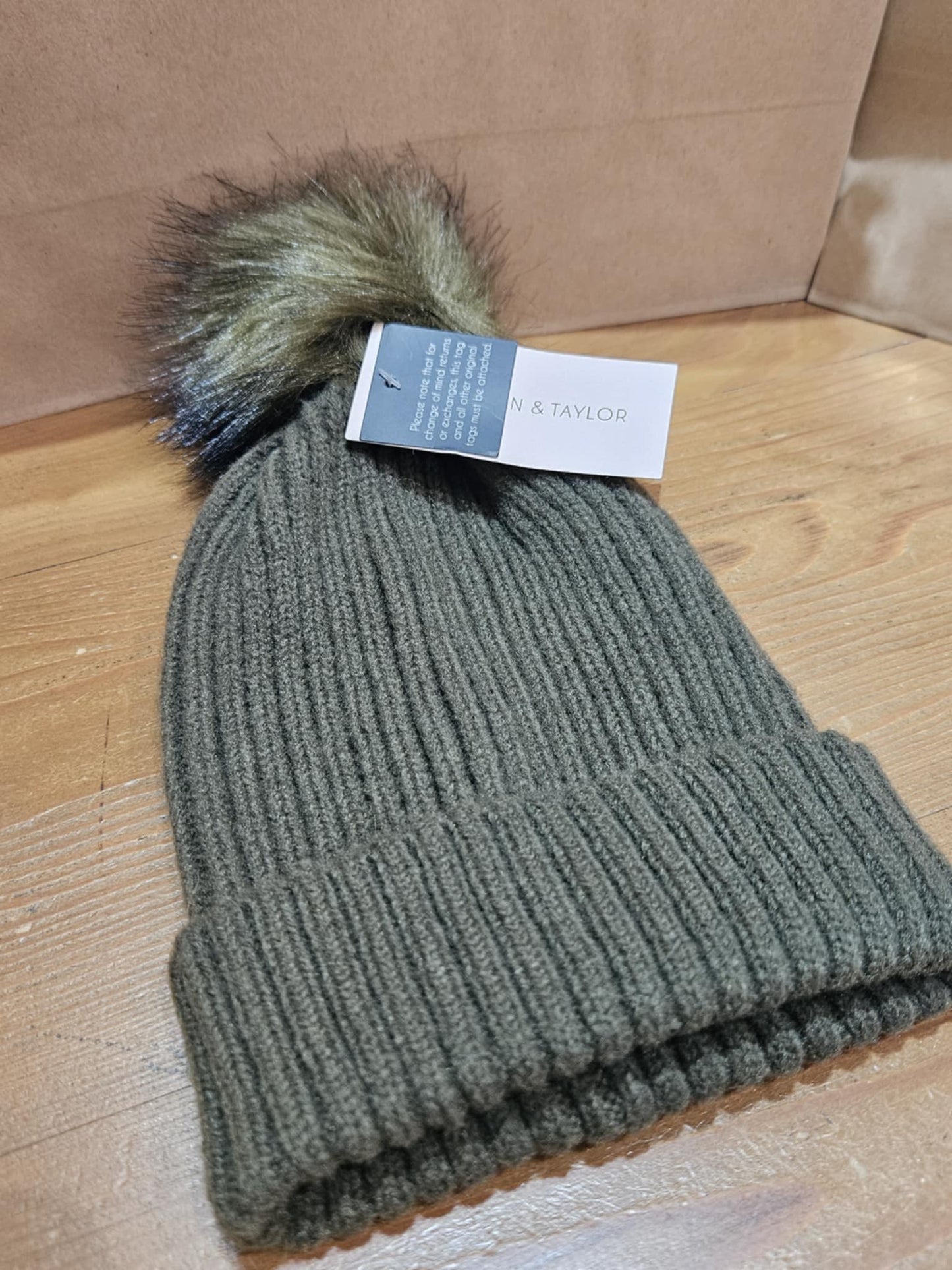 Khaki green Morgan & Taylor Zara Beanie with a faux fur pom-pom, laying flat on a wooden surface, showcasing its ribbed knit texture and cozy design.